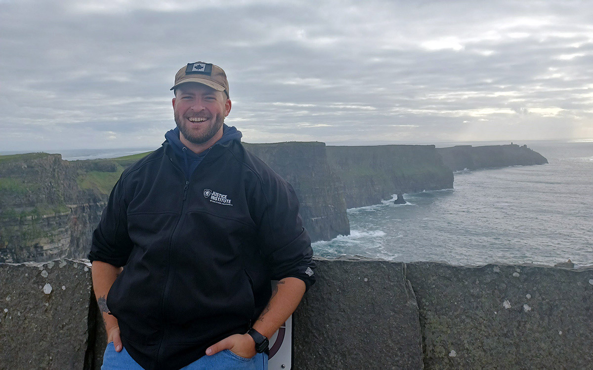 Man wearing hoodie and ball cap poses with view of cliffs and ocean in Ireland.