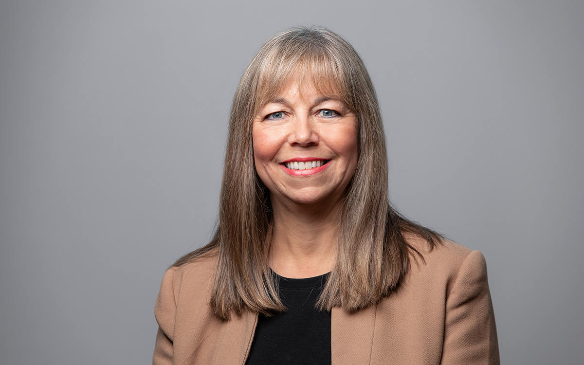 Woman wearing beige jacket over dark blouse against a grey background.