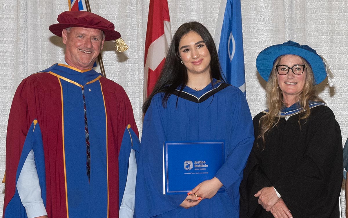 JIBC graduate in blue graduation gown poses between JIBC President and CEO Michel Tarko and 2023 honorary degree recipient Karen Fry.