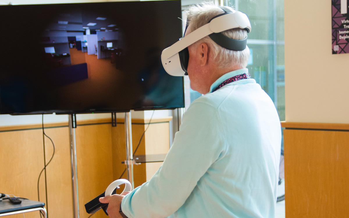 Man wearing virtual reality headset holds prop handgun in front of video screen showing training simulator.
