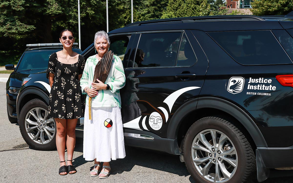 Musqueam artist Kamryn Sparrow and JIBC Elder-in-Residence Caroline Buckshot with JIBC Police Academy vehicle adorned with Sparrow's Indigenous design.