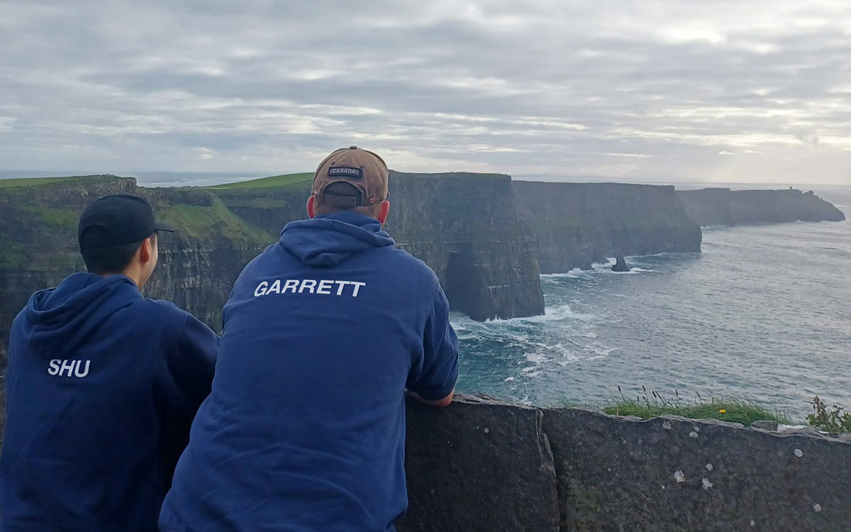 Two male students wearing hoodies and ball caps face away from the camera, looking at the view of cliffs and the ocean.