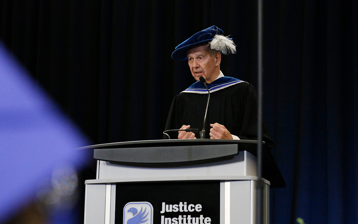 Man in black graduation robe and blue doctorate’s cap speaks at podium.