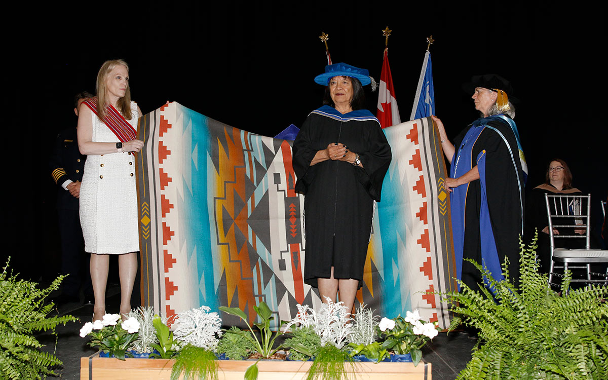 Two women hold up an Indigenous blanket while a woman in black robe and blue doctorate’s cap stands in the centre of it.
