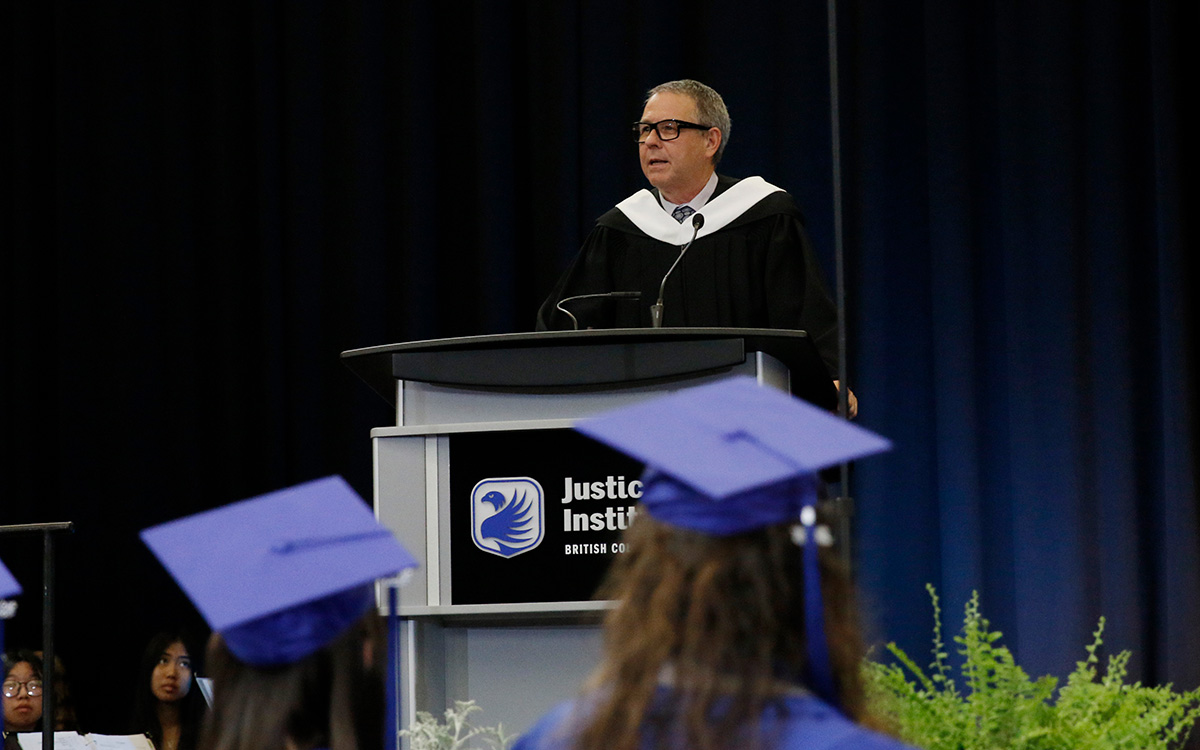 Man in glasses and wearing black graduation robe speaks at podium.