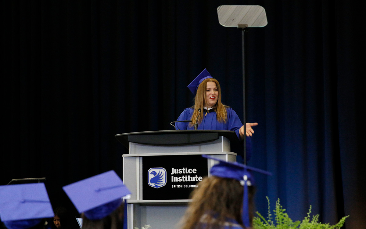 Woman in blue graduation gown and cap gestures with hand while speaking at a podium.