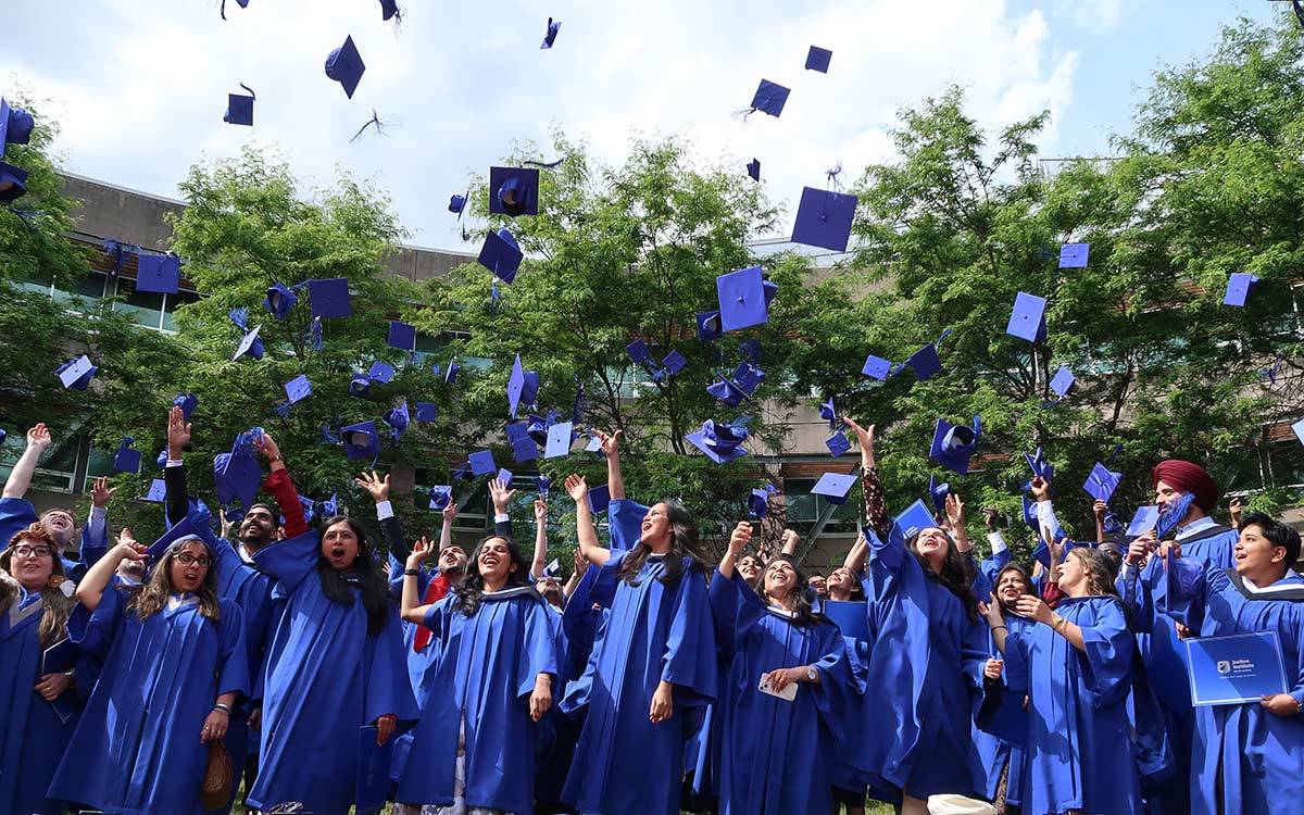 JIBC graduates in blue graduating gowns toss their grad caps in the air.