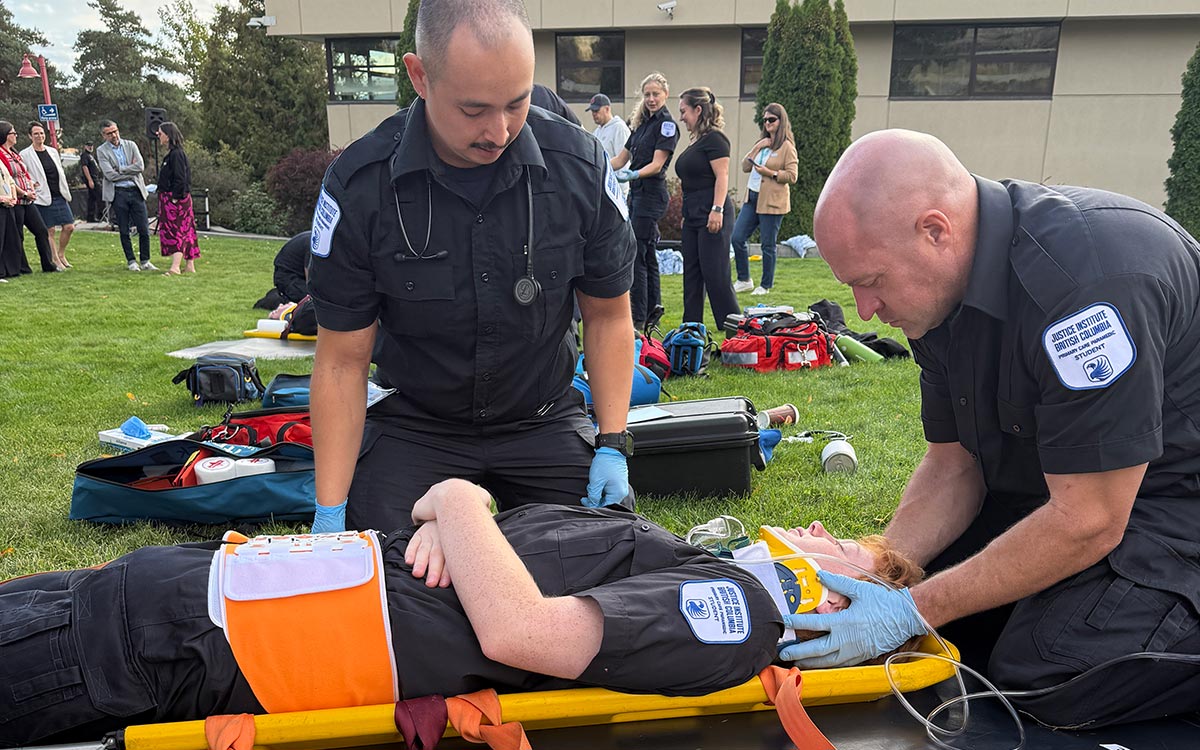 Two paramedic students in black uniforms secure a neck brace onto a third student on a stretcher.