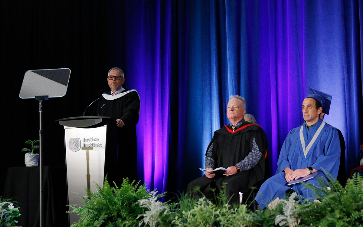 Man at podium speaks while two seated men look on.