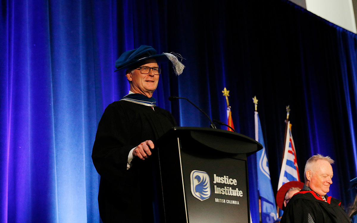 Man in black graduation gown and blue puffy hat speaks at podium.