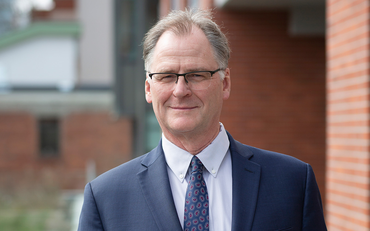 Man wearing glasses in blue suit and tie standing next to brick wall.
