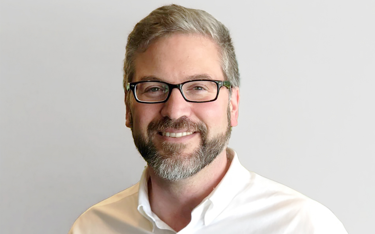 Head and shoulders of man with glasses and beard wearing white shirt.
