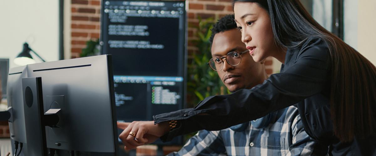 Standing woman points at computer monitor while seated man looks on.