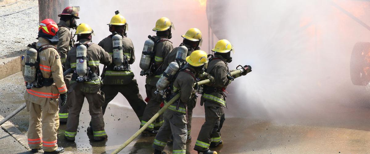 Group of firefighting students fighting fire while instructors look on.