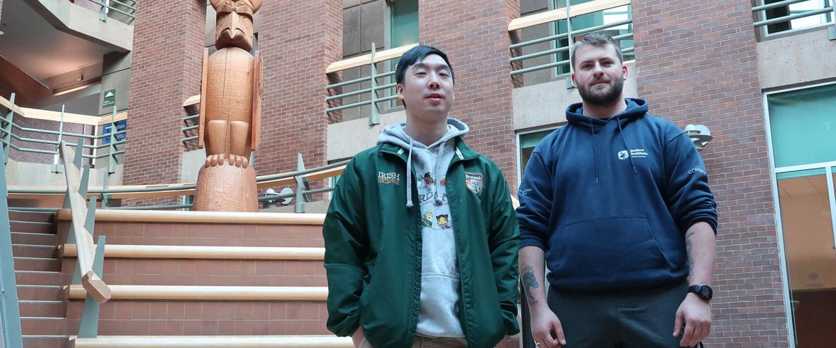 Two male students in atrium at JIBC's New Westminster campus, with Welcome Post in the background.