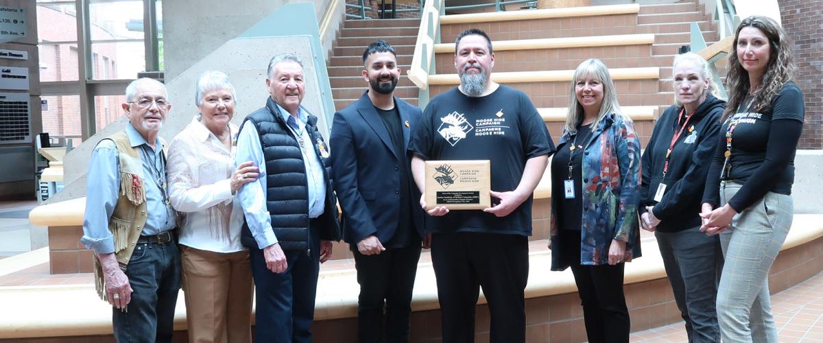 Eight people standing in a row with one man in a black Moose Hide Campaign t-shirt holding a plaque.