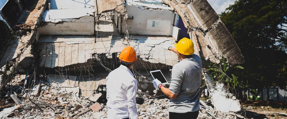 Two relief workers wearing hard hats look at collapsed building.