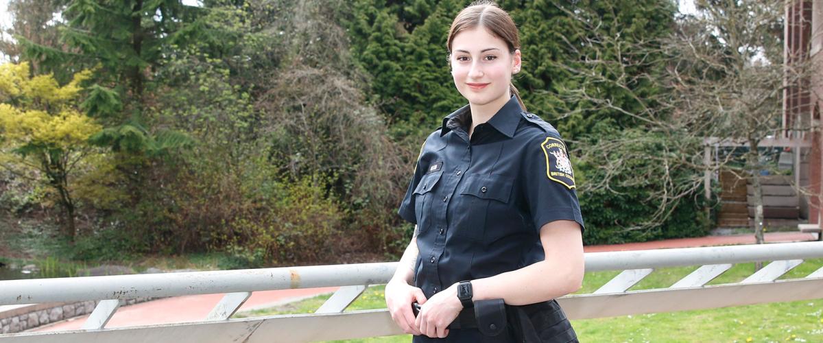 Woman in BC Corrections officer uniform standing at overlooking JIBC courtyard with hands resting on utility belt.