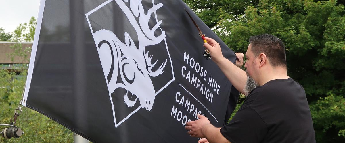 Man waves feather over a black flag with an image of a moose that says, "Moose Hide Campaign"