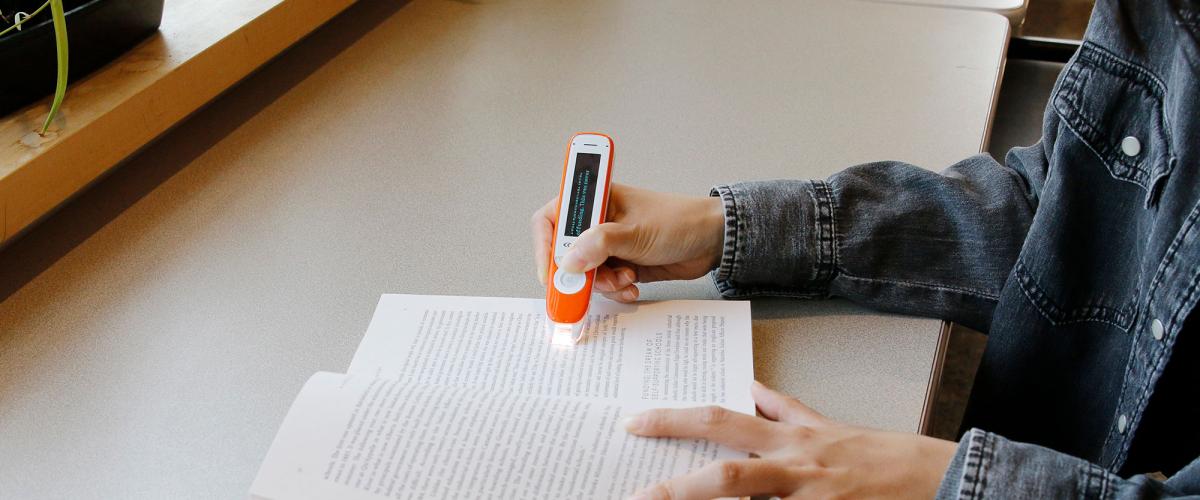 Close up of hands using a C-Pen device to highlight words in a book which it reads aloud.