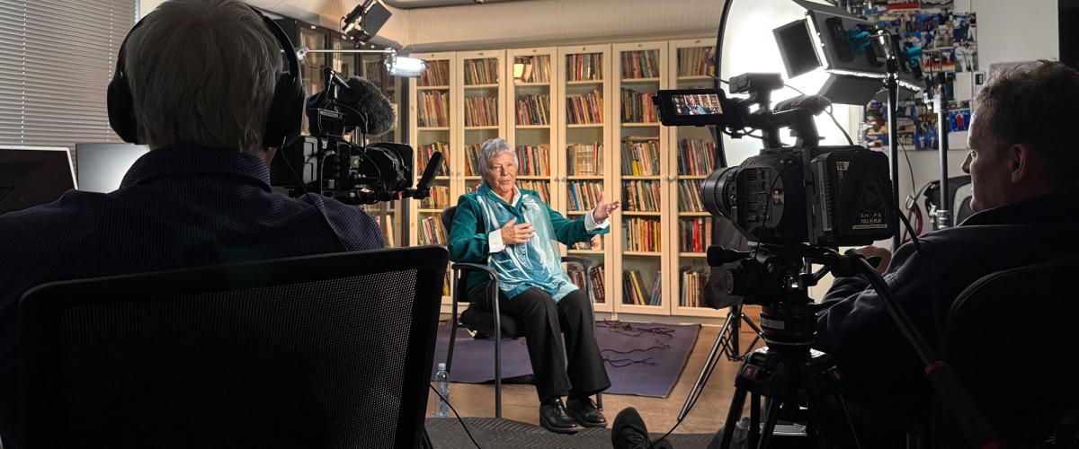 Woman sits in chair gesturing as two men sit behind lights and video cameras pointed at her.n