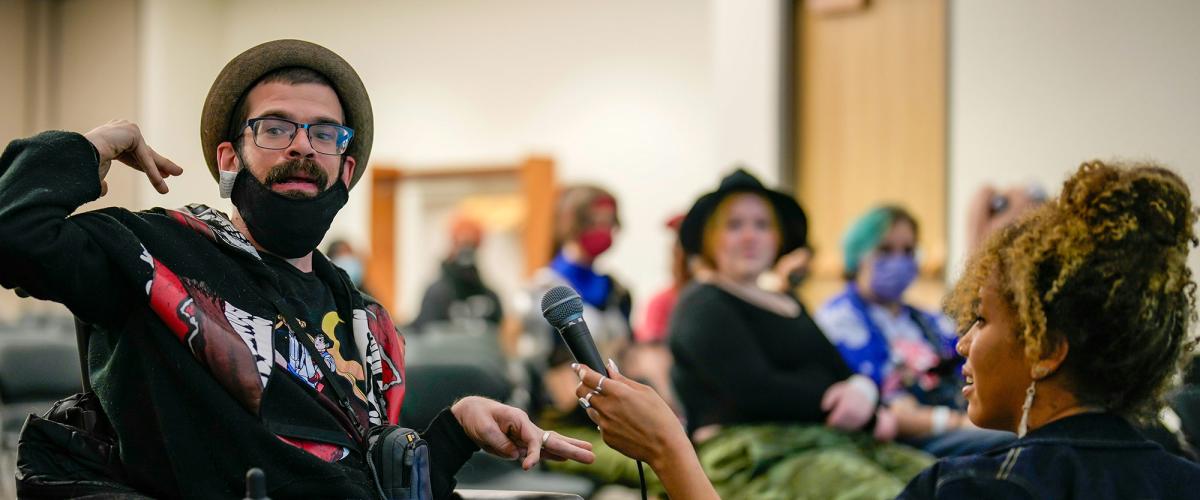 Man wearing a hat and glasses sits in a wheelchair and gestures while a woman holds a microphone in front of him.