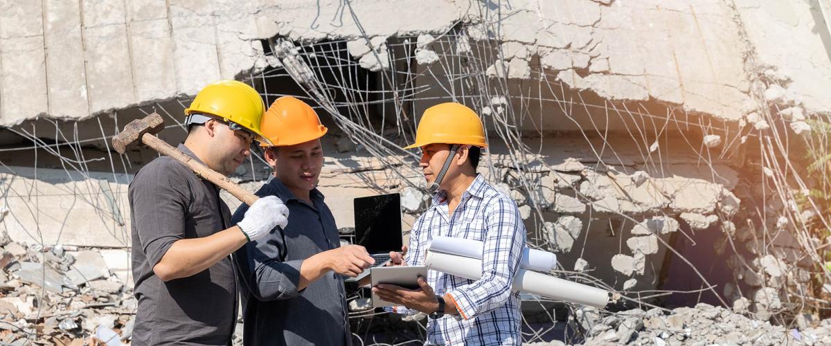 Three men in yellow hard hats look at a tablet while standing in front of a collapsed building.