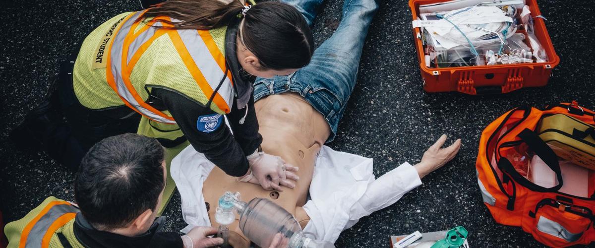 Two paramedic students in yellow reflective vests perform CPR on a dummy laying on the pavement.