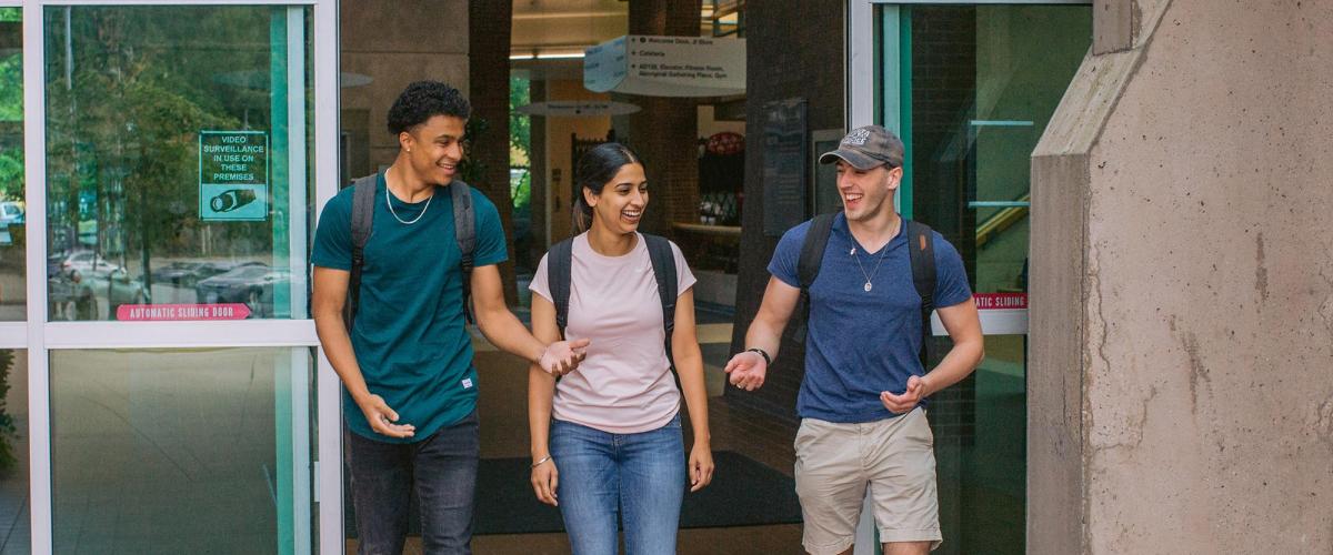 One female student walks out of campus doors flanked by two male students, all smiling in conversation.