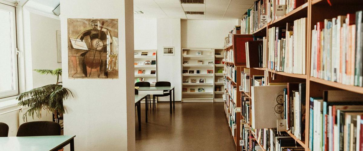 Room with bookshelves on right and tables and chairs by windows on left. 