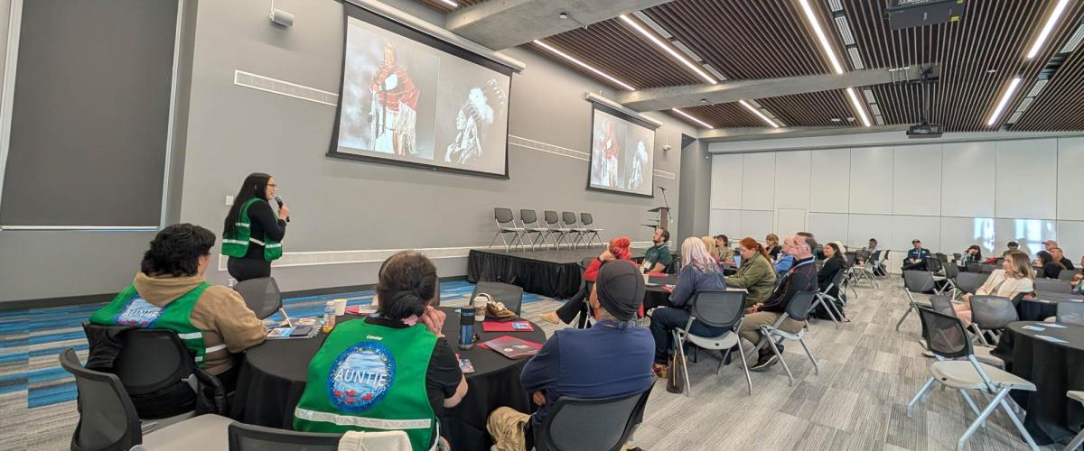 Large room with people in groups at tables and a person speaking at front..