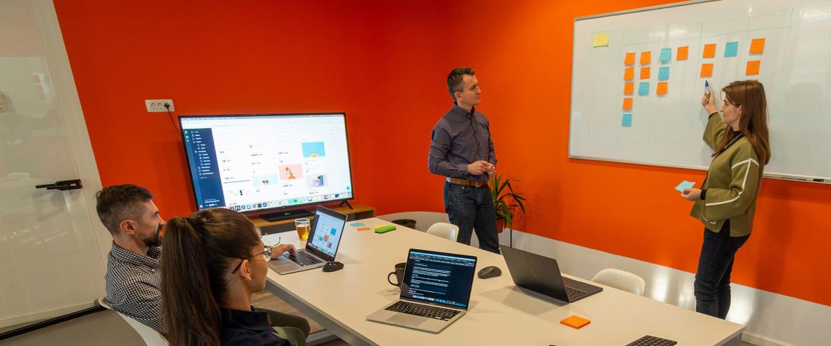 Two people seated watching two people at whiteboard with sticky notes on it.