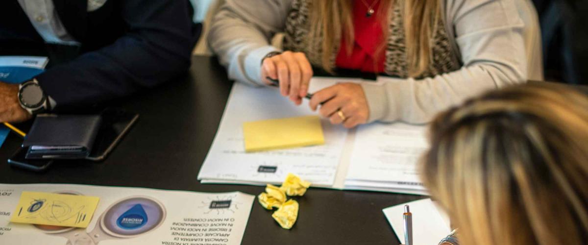 People seated around a table reviewing printed documents and yellow sticky notes, with a smartphone and colored stickers nearby as one person writes with a pen.