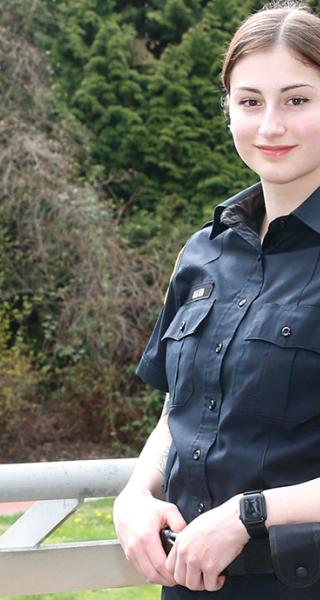 Woman in BC Corrections officer uniform standing at overlooking JIBC courtyard with hands resting on utility belt.