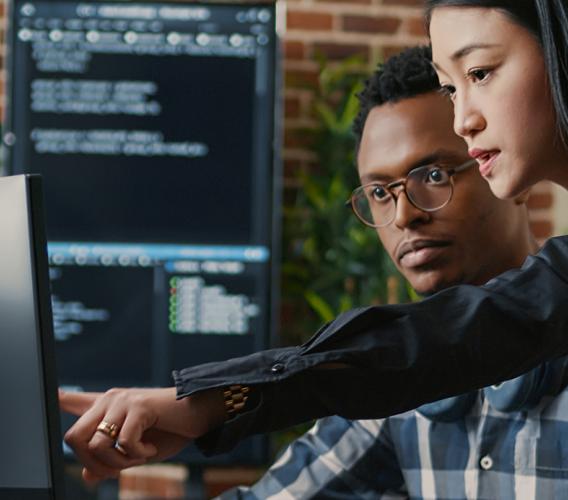 Standing woman points at computer monitor while seated man looks on.