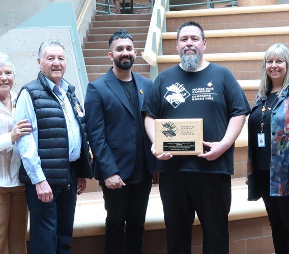 Eight people standing in a row with one man in a black Moose Hide Campaign t-shirt holding a plaque.