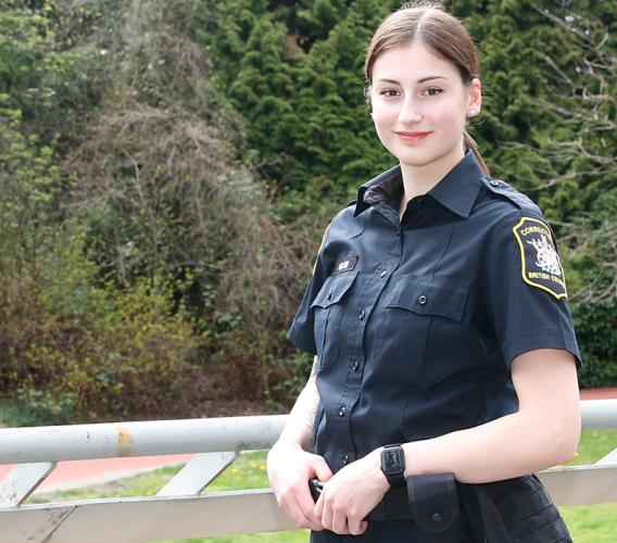 Woman in BC Corrections officer uniform standing at overlooking JIBC courtyard with hands resting on utility belt.