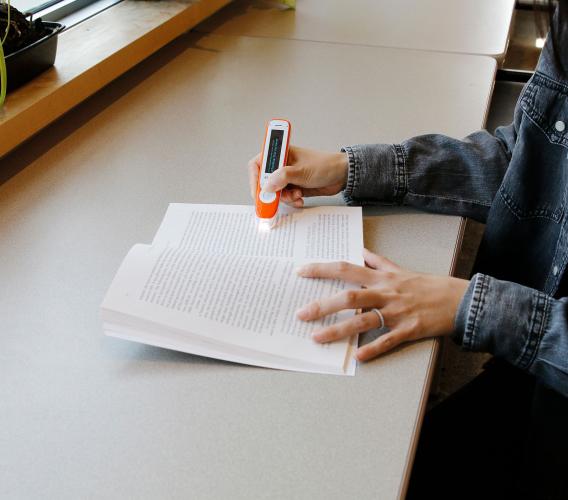 Close up of hands using a C-Pen device to highlight words in a book which it reads aloud.