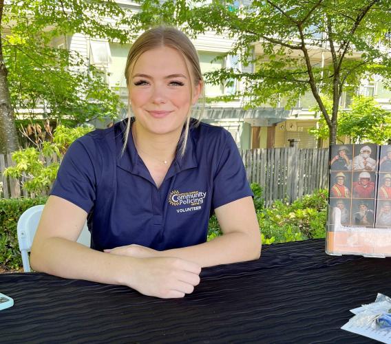 Woman sits at a table outdoors wearing golf shirt that says "community policing volunteer."