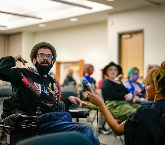 Man wearing a hat and glasses sits in a wheelchair and gestures while a woman holds a microphone in front of him.
