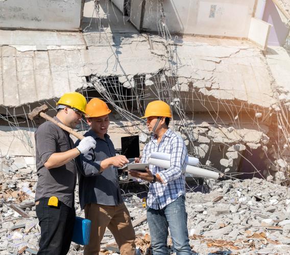 Three men in yellow hard hats look at a tablet while standing in front of a collapsed building.
