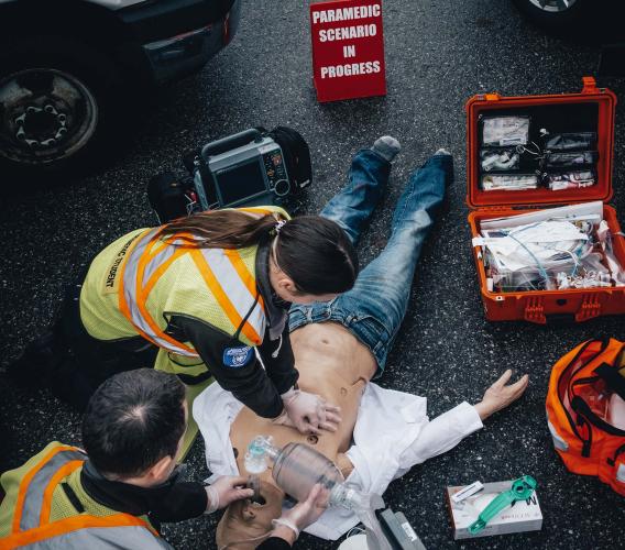 Two paramedic students in yellow reflective vests perform CPR on a dummy laying on the pavement.
