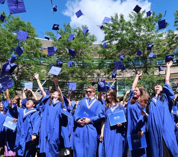 JIBC graduates in blue convocation gowns toss their caps in the air.