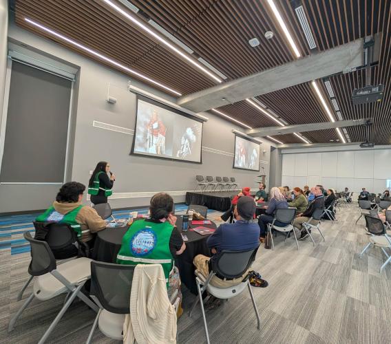 Large room with people in groups at tables and a person speaking at front..