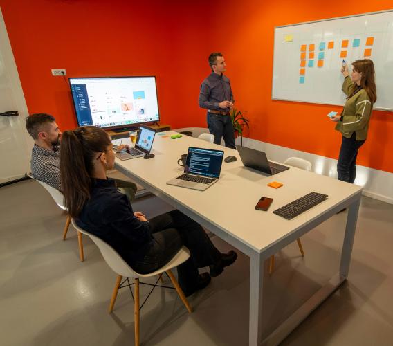 Two people seated watching two people at whiteboard with sticky notes on it.