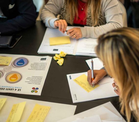 People seated around a table reviewing printed documents and yellow sticky notes, with a smartphone and colored stickers nearby as one person writes with a pen.