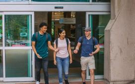 One female student walks out of campus doors flanked by two male students, all smiling in conversation.