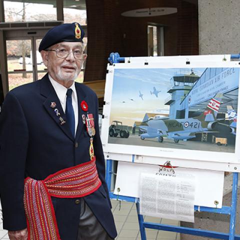 Elder Ken Pruden wearing Royal Canadian Air Force uniform and Metis sash stands next to poster depicting an aircraft hangar.