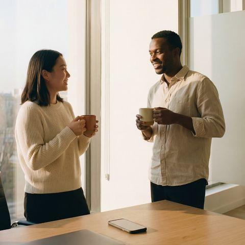 Two people smiling and holding mugs while standing near a window by a wooden table with a smartphone, in a bright modern room.