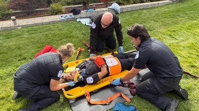 Three paramedic students in black uniforms secure a fourth student onto a stretcher.